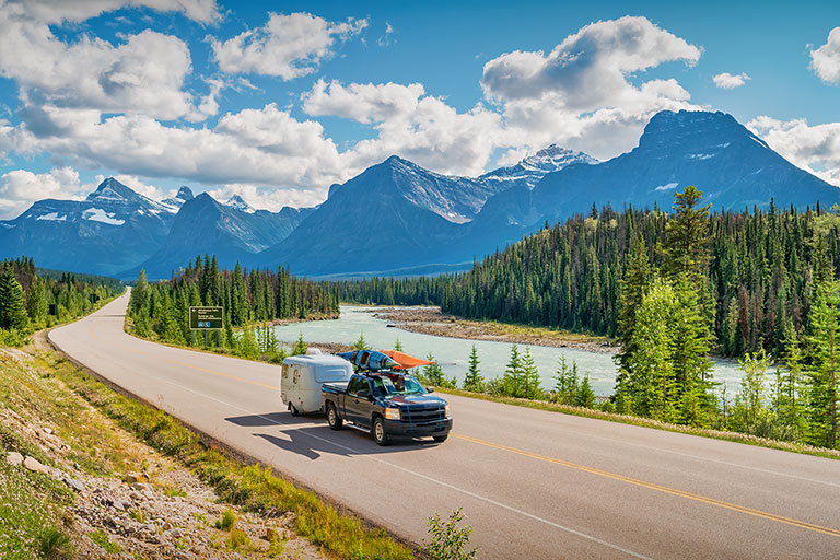 Image of a truck with a trailer and kayaks driving down a road with a lake and mountains.